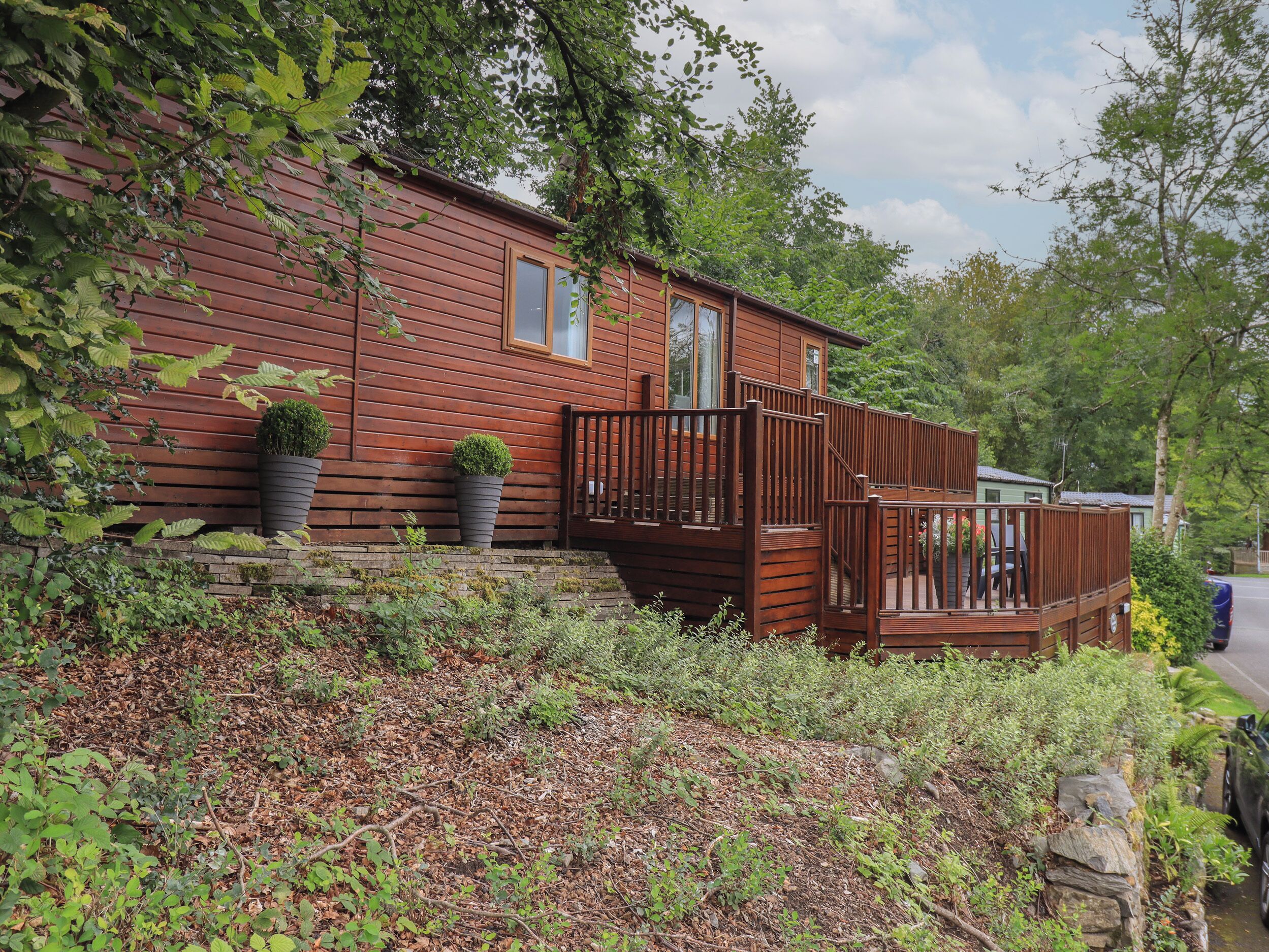 A wooden cabin with a deck, surrounded by dense foliage and a stone wall.