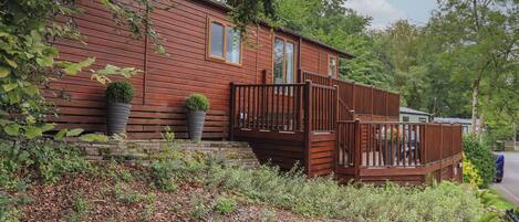 A wooden cabin with a deck, surrounded by dense foliage and a stone wall.