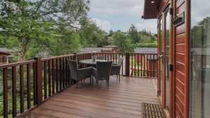 Une terrasse en bois, agrémentée d’une table et de chaises, entourée d’une balustrade en bois, et offrant une vue sur un jardin et d’autres bâtiments.