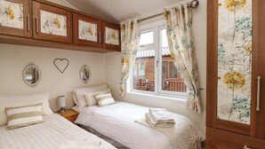 A bedroom with two beds, wooden cabinets with floral designs, a window with curtains, and a chandelier. - Shoreside Lodge (Windermere)