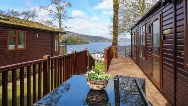 A wooden deck with a glass table and a basket of flowers, leading to a cabin with a view of a lake. - Shoreside Lodge (Windermere)