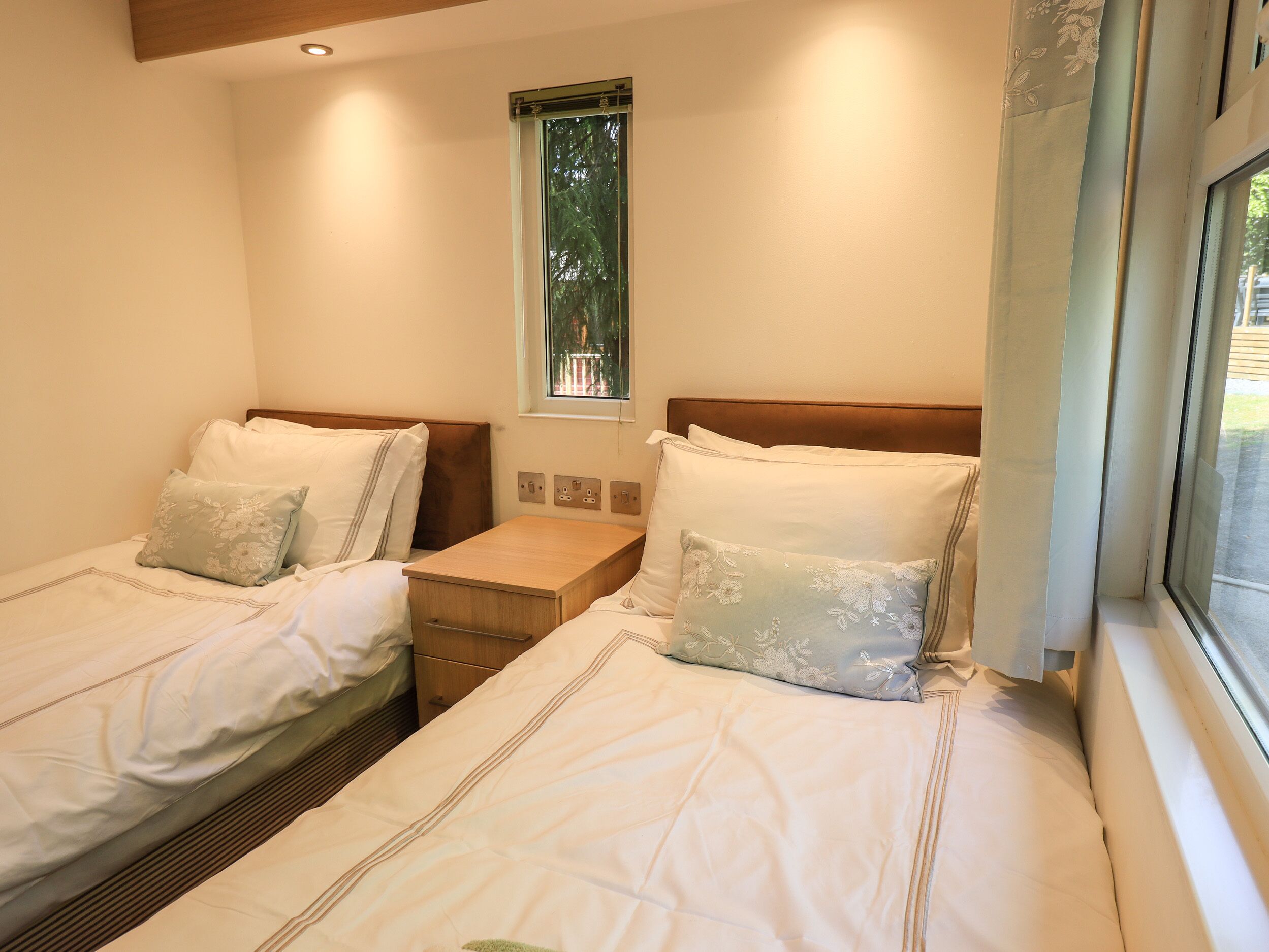 Un dormitorio con dos camas individuales, una mesita de noche entre ellas, una ventana con vistas a árboles y un enchufe en la pared.