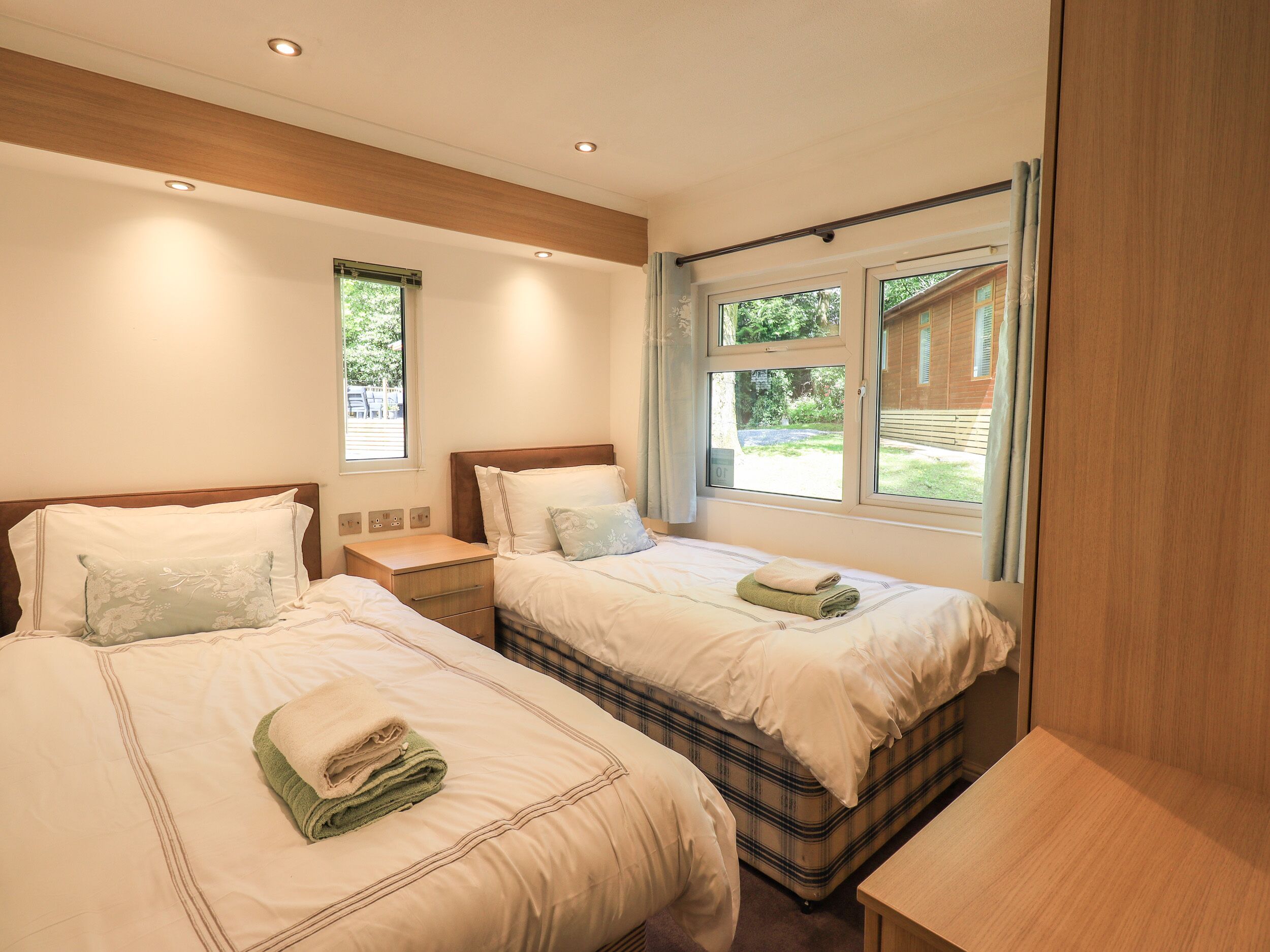 A hotel room with two beds, a wooden headboard, a small bedside table, and a window with a view of greenery.