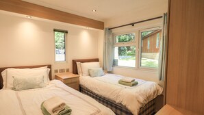 A hotel room with two beds, a wooden headboard, a small bedside table, and a window with a view of greenery. - Forest Pines Lodge (Windermere)