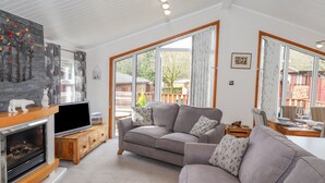 A cozy living room with a fireplace, a grey sofa, and large windows offering a view of the outdoors. - Limefitt Lodge (Windermere)