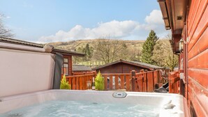 A hot tub with a wooden deck, a wooden fence, and a view of a cabin in the background. - Limefitt Lodge (Windermere)