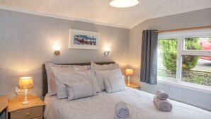 A neatly made bed with pillows and rolled towels, a bedside lamp, a framed picture on the wall, and a window with curtains. - Limefitt Lodge (Windermere)