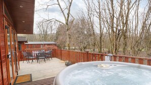 A wooden deck with a hot tub, outdoor seating, and a view of trees and a building in the background. - Wainwright Lodge (Windermere)