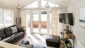 Una sala de estar moderna con un sofá de cuero, una mesa de centro de madera y un televisor. Hay una puerta corrediza de vidrio que da a un área exterior con una terraza de madera.