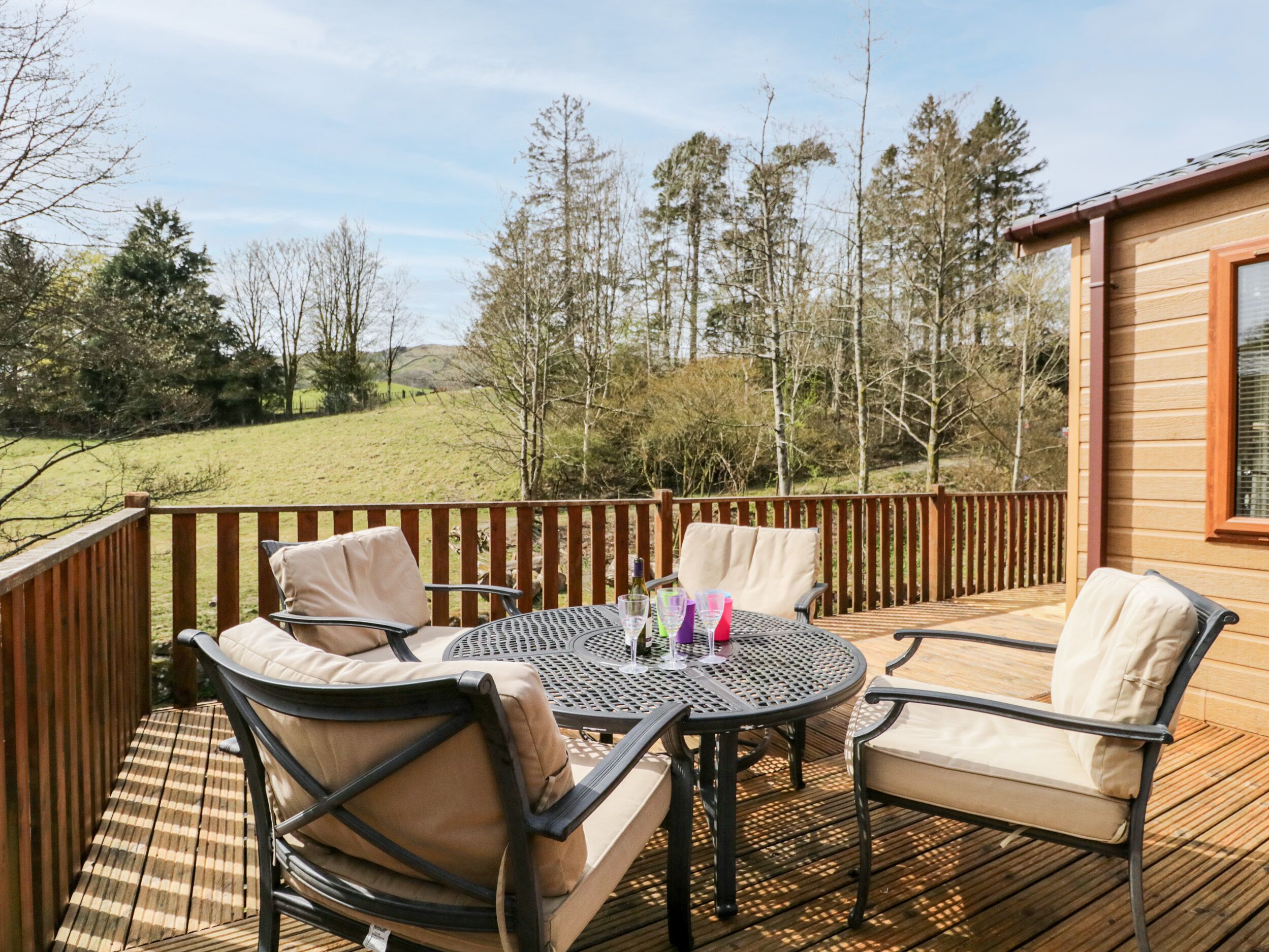 A wooden deck with outdoor furniture, including chairs and a round table, overlooking a grassy landscape with trees.