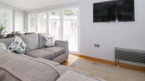 Una sala de estar moderna con un sofá gris, un televisor de pantalla plana montado en la pared y ventanas grandes con persianas.