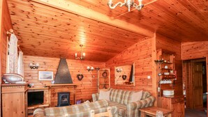 Ein gemütliches Blockhaus-Interieur mit Kamin, kariertem Sofa, Esstisch für zwei Personen und Holzregalen mit diversen Gegenständen.