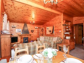 Interior acogedor de una cabaña de madera con chimenea, un sofá de cuadros, una mesa de comedor para dos personas y estanterías de madera con diversos objetos.