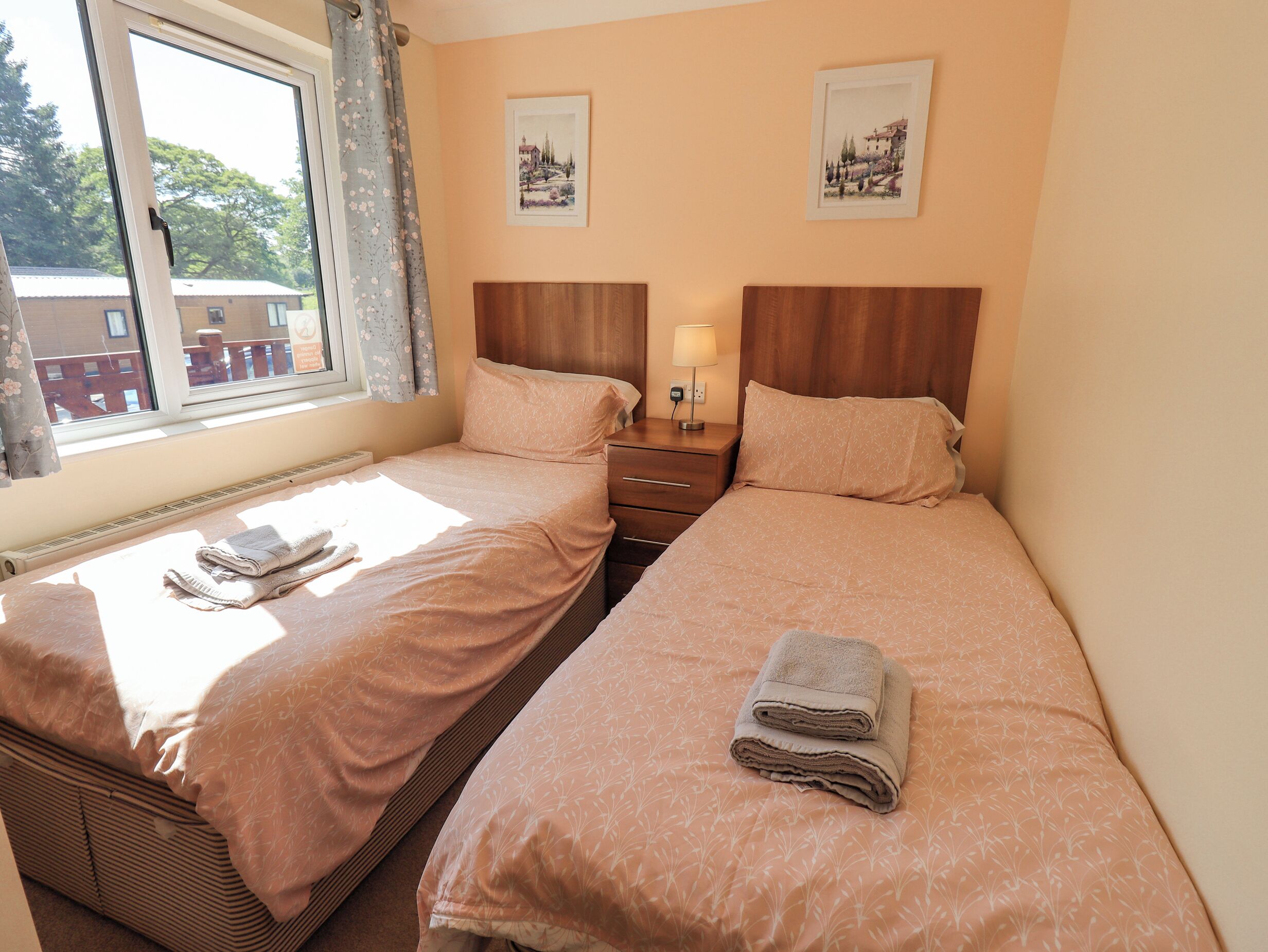 A double room with two beds, a window with curtains, a bedside table with a lamp, and two framed pictures on the wall.