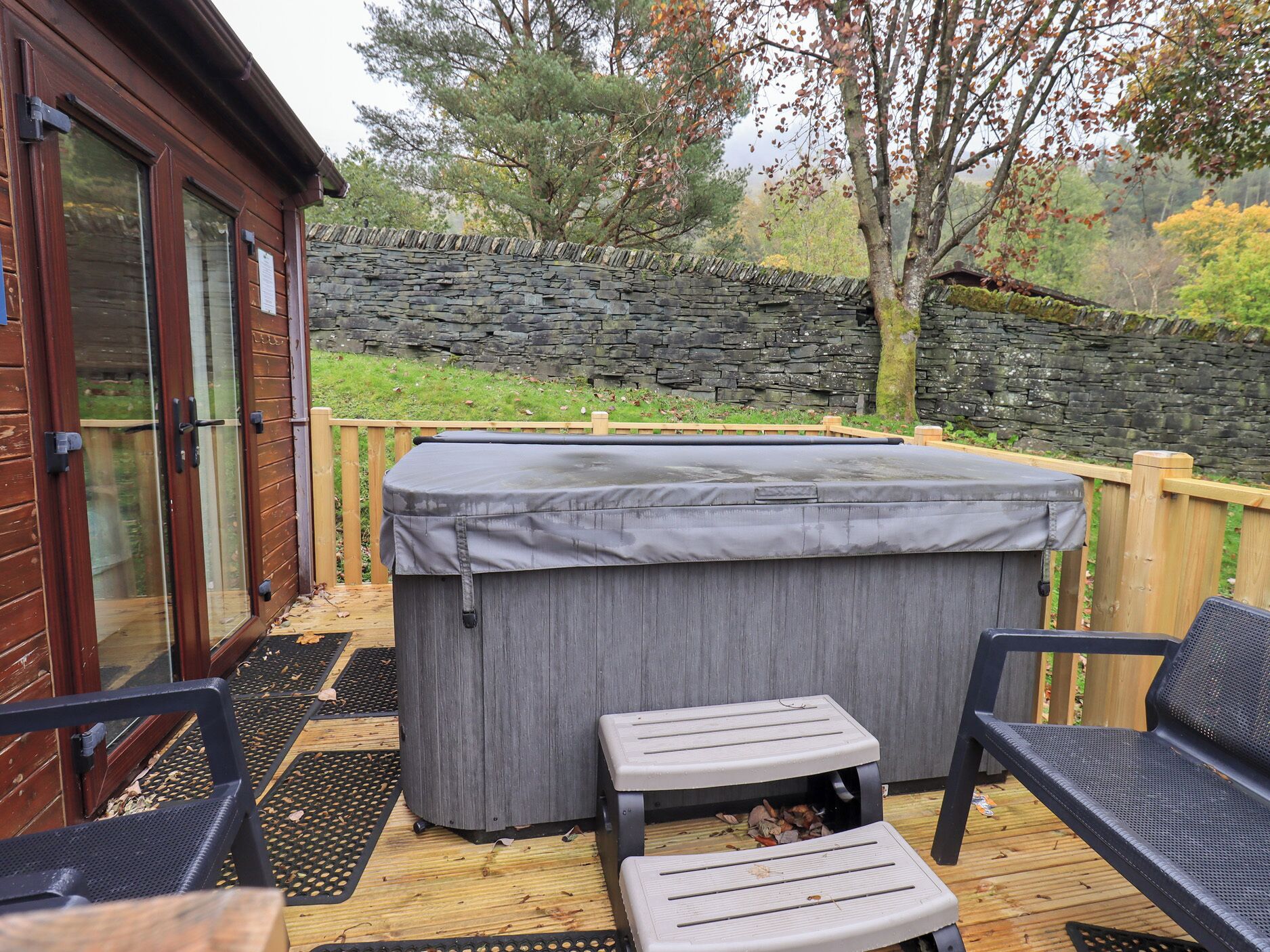 A wooden deck with a stone wall, a covered hot tub, and two outdoor chairs.