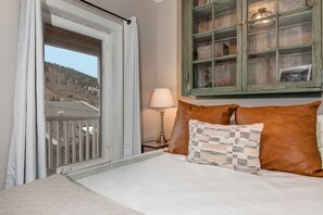 Relax in a spacious bedroom with a large bed, featuring a quilted headboard and two pillows, and enjoy a view of the surrounding mountains from the balcony.