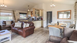 A cozy living room with a brown leather sofa, a dining table with chairs, a kitchen area with cabinets and appliances, and a large mirror on the wall. - Sunny Brow Lodge (Windermere)