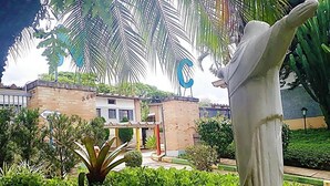 Um edifício com um grande letreiro azul em formato de 'C', rodeado por vegetação exuberante e uma escultura branca imponente.