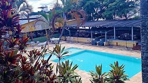 Uma piscina com deck ladrilhado, rodeada por plantas tropicais e uma área de estar coberta.