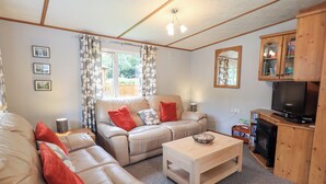Ein gemütliches Wohnzimmer mit einer Couch, einem Couchtisch und einer TV-Konsole. An der Wand hängen gerahmte Bilder und es gibt ein Fenster mit Vorhängen.