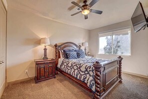 Un dormitorio con cama de madera, dos mesitas de noche, ventilador de techo, ventana y televisor montado en la pared.