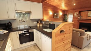 A modern kitchen with white cabinets, a black countertop, and stainless steel appliances. There is a wooden dining area with a sofa and a fireplace. - The Fold (Keswick)