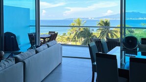 A modern living room with a large glass wall offering a view of the ocean and palm trees. - Amazing Jewel Beach front Condo with great views (Bucerias/ Nuevo Vallarta)