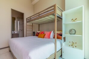 Una cama litera con cabecera blanca, una estantería blanca con objetos decorativos, y un baño con espejo y lavabo a la vista.