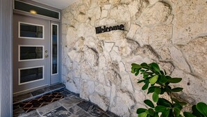 Un muro de piedra con un letrero que dice 'Welcome', una puerta gris con paneles de vidrio esmerilado y un felpudo.