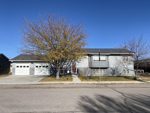 A two-story residential building with a garage, a tree in front, and a clear blue sky. - Montana Home close to golfing, fishing, skiing, hiking, hunting and downtown (Dillon)