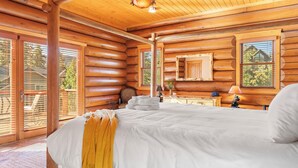 A log cabin bedroom with a large bed, wooden walls, and a window overlooking a deck.
