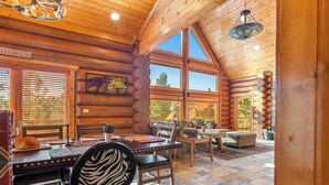 A log cabin interior with a dining area, large windows, and a cozy seating arrangement.