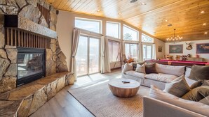 Amplia sala de estar con chimenea de piedra, techo de madera, ventanas grandes y un rincón de estar acogedor.