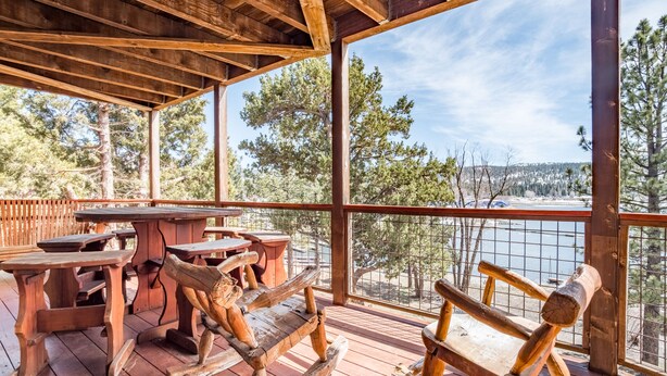 Una terraza de madera con mobiliario rústico, que incluye bancos y una mesa, con vistas a un paisaje pintoresco con árboles y una masa de agua.