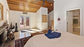 A spacious room with a high ceiling, wooden beams, a ceiling fan, a bed with a blue blanket, a fireplace, and a large window. - Views at Wolf Lodge by AvantStay | Magnificent Views w/ Hot Tub, Sauna, Games (Big Bear Lake)