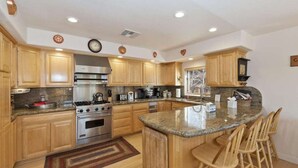 A kitchen with wooden cabinets, granite countertops, stainless steel appliances, and a central island with bar stools. - Views at Wolf Lodge by AvantStay | Magnificent Views w/ Hot Tub, Sauna, Games (Big Bear Lake)