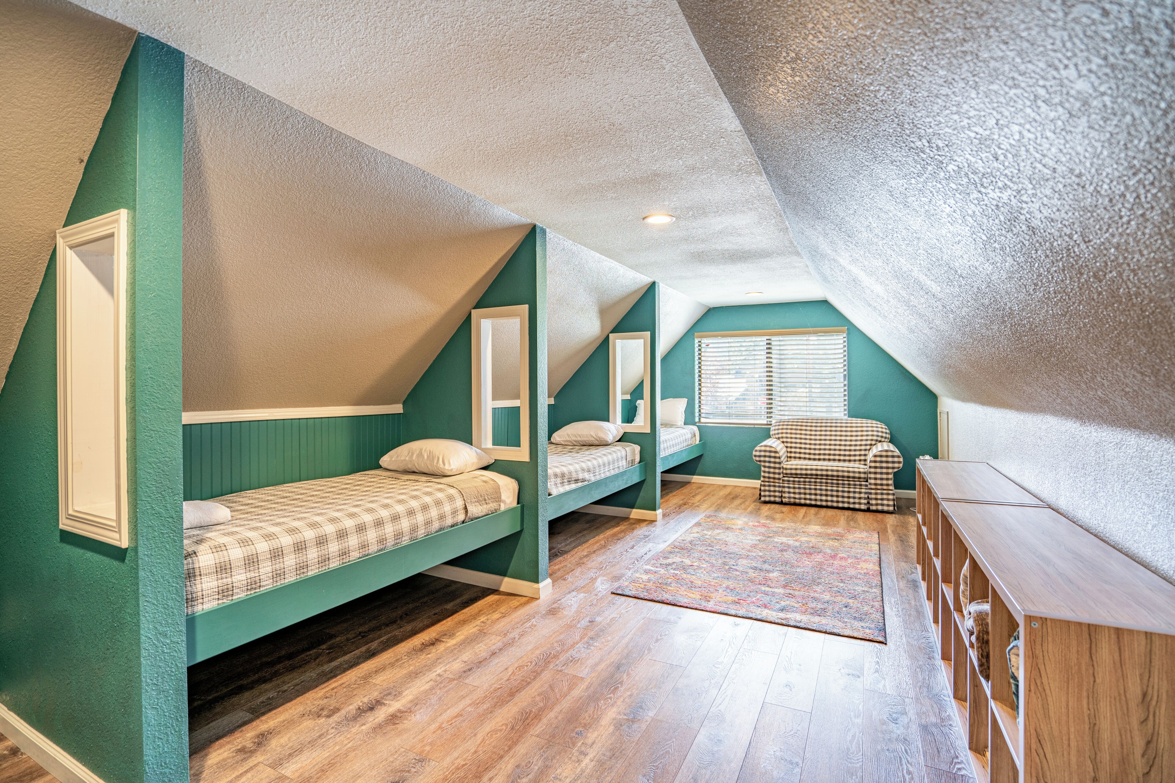A room with a sloped ceiling, two bunk beds with plaid bedding, a checkered chair, and a wooden bench.
