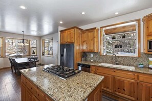 A kitchen with granite countertops, wooden cabinets, a stainless steel refrigerator, and a window with a view of trees.