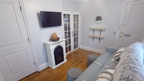 Een woonkamer met een open haard, een aan de muur bevestigde televisie, een bank en een klein, rond poef.