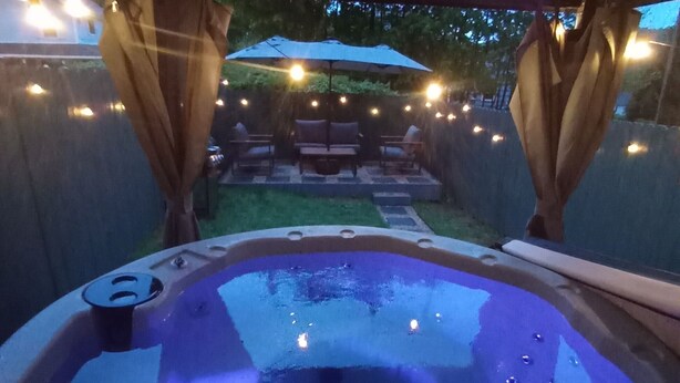 Een hottub onder een pergola met lichtjes, omringd door een terras met stoelen en een parasol.