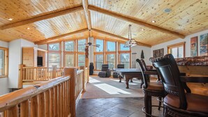 A spacious room with a high ceiling, wooden beams, large windows, a pool table, and leather chairs.