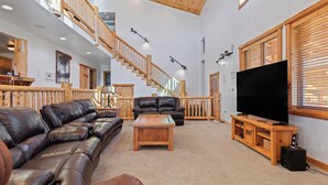 A spacious living room with a high ceiling, wooden staircase, leather sofas, a wooden coffee table, and a large flat-screen TV.