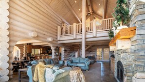 Un intérieur spacieux de chalet en bois, avec un plafond haut, une cheminée en pierre et un balcon donnant sur l’espace de vie.
