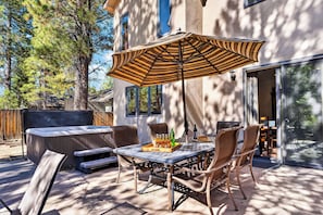 An outdoor patio with a table, chairs, a large striped umbrella, and a hot tub. - Walk to Lake | 6BR Cabin w/ Game Room, Hot Tub | Big Bear Castle (Big Bear Lake)