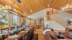A spacious living area with a wooden ceiling, a dining table set for a meal, a staircase, and a kitchen in the background.