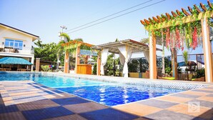 Une piscine avec un revêtement de sol carrelé, un espace détente couvert agrémenté d’une pergola, et un bâtiment avec un balcon en arrière-plan.