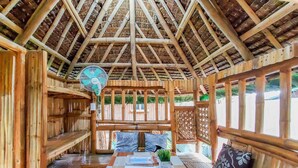 Un intérieur en bois, avec un toit de chaume, un ventilateur et une table recouverte d’une nappe à carreaux.