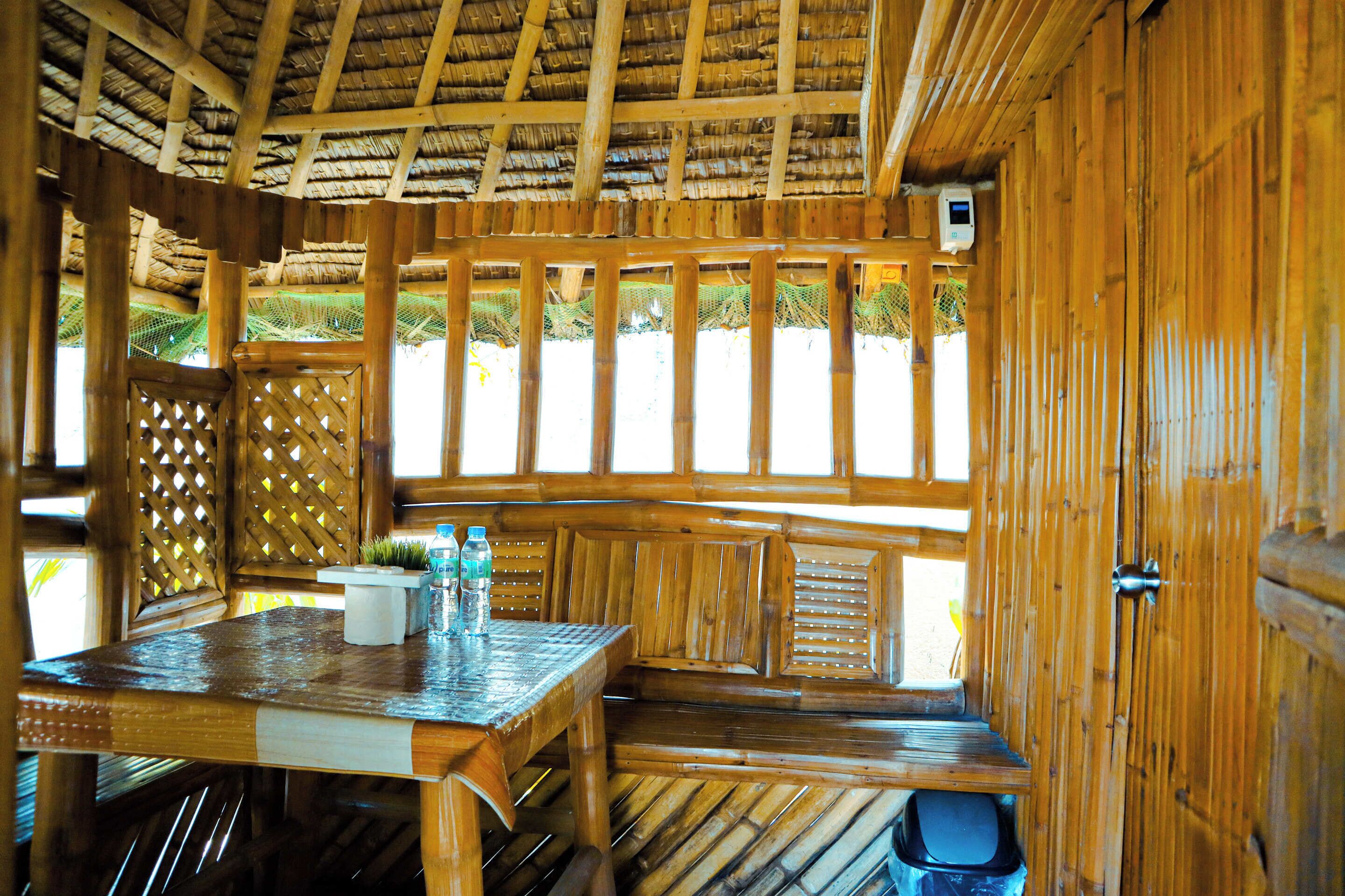 A wooden interior with a thatched roof, a wooden bench, and a table with water bottles.
