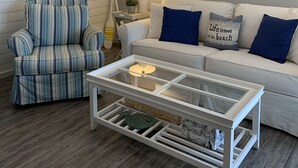 Un salón con un sillón a rayas, un sofá blanco con cojines azules, una mesa de centro de cristal y un cuadro de una playa.