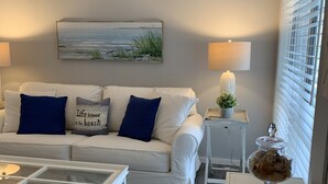 Un salón con un sofá blanco, cojines azules, una mesa de centro de vidrio, una mesita auxiliar con una lámpara, un cuenco decorativo y un cuadro en la pared.
